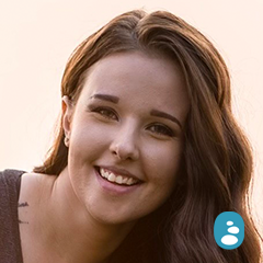A person with long brown hair and glowing skin smiles warmly at the camera. A small blue speech bubble icon is displayed in the lower right corner of the image. - OYESPA Aveda Lifestyle | nver Grove Heights, MN