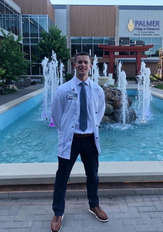 A person in a white lab coat stands in front of a beautiful fountain outside the Palmer College of Chiropractic. - OYESPA Aveda Lifestyle | nver Grove Heights, MN