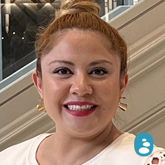 A person with a bun hairstyle and wearing a white shirt is smiling. There are stairs and a reflective surface in the background, hinting at the sleek design cherished by the oyespa team. A blue icon is at the bottom right corner of the image. - OYESPA Aveda Lifestyle | nver Grove Heights, MN