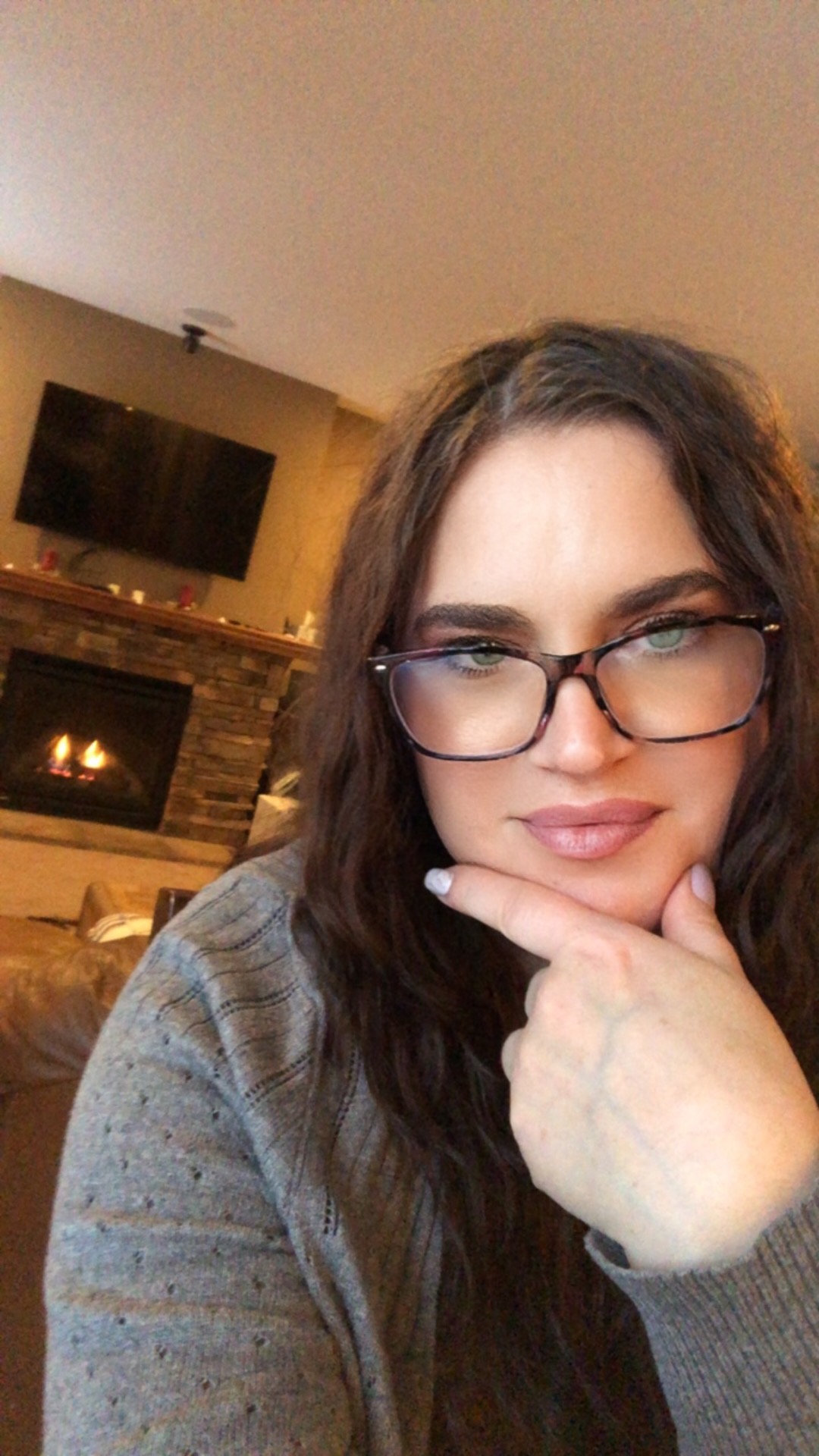 A person with long hair and glasses sits indoors, resting their chin on their hand. A fireplace and television are visible in the background. - OYESPA Aveda Lifestyle | nver Grove Heights, MN