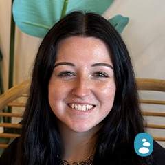 A person with long dark hair smiling, seated on a wooden chair with green leaves in the background. A small blue icon is at the bottom right corner. - OYESPA Aveda Lifestyle | nver Grove Heights, MN