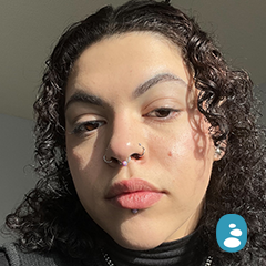 Person with curly hair, nose, and lip piercings, looking at the camera, with sunlight on their face. - OYESPA Aveda Lifestyle | nver Grove Heights, MN