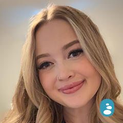 A young woman with long, wavy blonde hair smiles at the camera against a neutral background. A small blue icon with a white person symbol is in the bottom right corner. - OYESPA Aveda Lifestyle | nver Grove Heights, MN