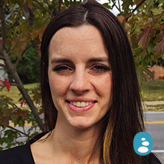 A woman with long brown hair and a black top is standing outdoors in front of trees with green and red leaves. - OYESPA Aveda Lifestyle | nver Grove Heights, MN