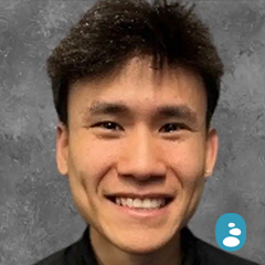 A person with short, dark hair smiling at the camera in front of a gray, textured background. - OYESPA Aveda Lifestyle | nver Grove Heights, MN