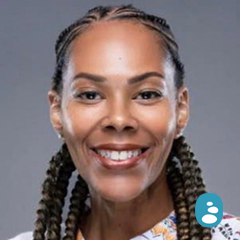 A woman with braided hair smiles at the camera against a plain grey background. A small blue icon with a person symbol appears in the lower right corner. - OYESPA Aveda Lifestyle | nver Grove Heights, MN