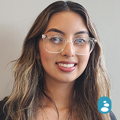 Woman with long, wavy brown hair and clear glasses smiling at the camera against a plain light background. Blue user icon in bottom right corner. - OYESPA Aveda Lifestyle | nver Grove Heights, MN