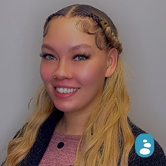A woman with long, wavy blonde hair and braids smiles at the camera against a plain light-colored background. She is wearing a pink top and a dark sweater. - OYESPA Aveda Lifestyle | nver Grove Heights, MN