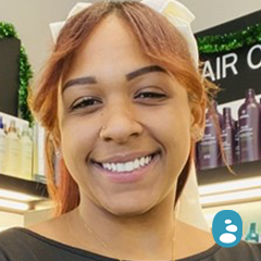 A person with reddish-brown hair and a white headband smiles in front of shelves with hair products. - OYESPA Aveda Lifestyle | nver Grove Heights, MN