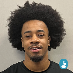 A man with a large afro hairstyle and a trimmed goatee smiles slightly at the camera, standing against a plain light-colored wall. A small blue icon is in the bottom right corner. - OYESPA Aveda Lifestyle | nver Grove Heights, MN
